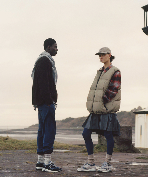 Knee down view of a man and woman wearing New Balance Made in UK sneakers standing on a concrete floor against a grey wall.