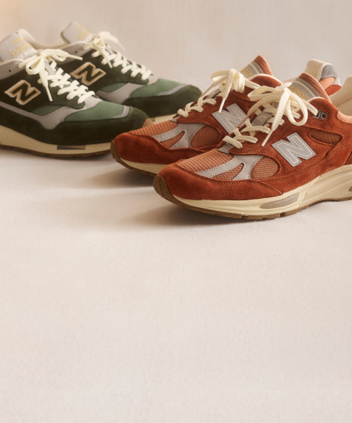 Knee down view of a man and woman wearing New Balance Made in UK sneakers standing on a concrete floor against a grey wall.