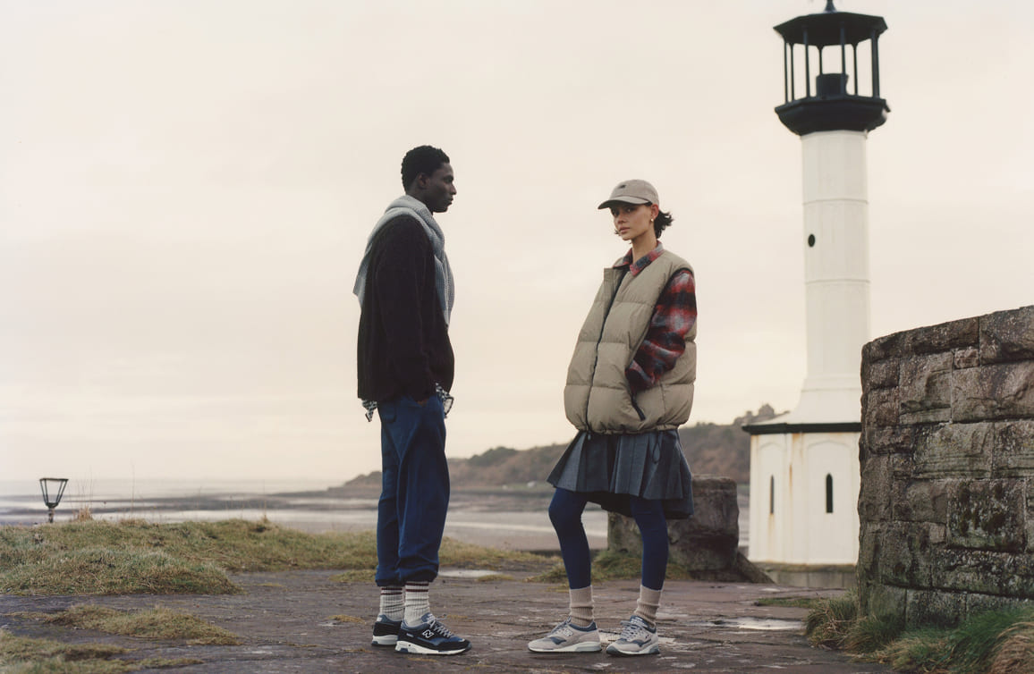 Knee down view of a man and woman wearing New Balance Made in UK sneakers standing on a concrete floor against a grey wall.
